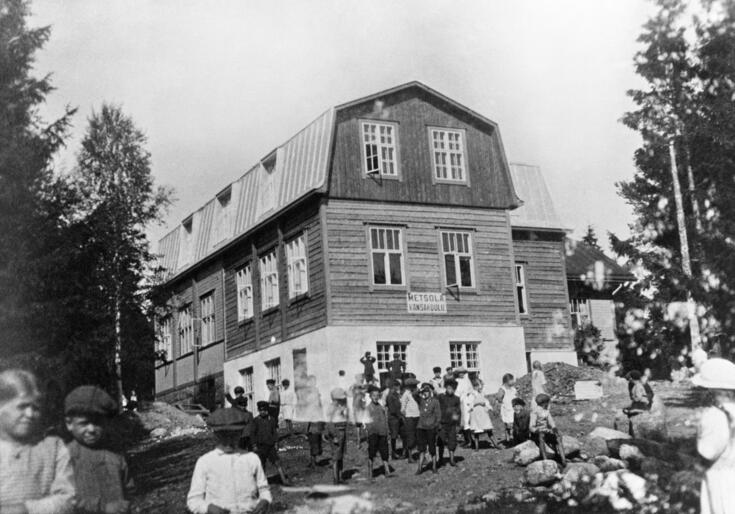 Keskellä koulurakennus, jonka seinässä kyltti Metsola kansakoulu. Etualalla lapsia, pojilla lakit päässä, tytöillä esiliinat.