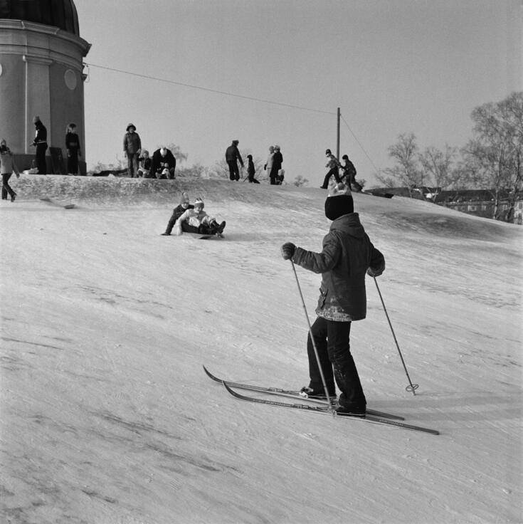 Children sledding in Kaivopuisto. In the foreground, a boy on skis with ski poles in his hands. In the background, two children ride together on a sled. The observatory stands in the background.