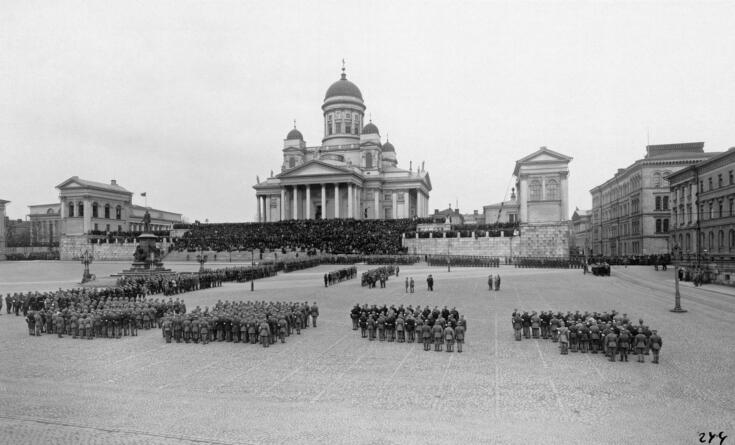 Jääkärit ovat järjestäytyneet paraatimuodostelmiin ympäri Senaatintoria. Ryhmittymien keskelle on kokoontunut oletettavasti jääkäriprikaatin päällystöä. Yleisö on kerääntynyt seuraamaan tapahtumia tuomiokirkon portaille.