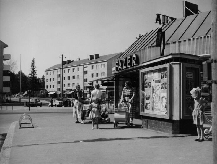 Utanför köpcentret går kvinnor och barn. En del av kvinnorna skuffar barnvagn. På sidan av byggnaden syns en glasvitrin med affischer i olika storlekar. På byggnadens väg finns Fazers skylt. I bakgrunden syns våningshus. 
