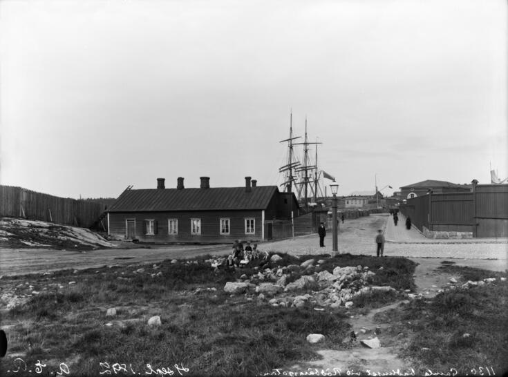 Gamla badinrättningens träbyggnad, i bakgrunden segelfartygsmaster, i förgrunden impediment och barn sittande på en sten. 