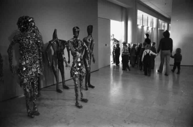 A lobby with a group of metallic, robotic, jagged sculptures of human height. Children can be seen walking out the door in the background.