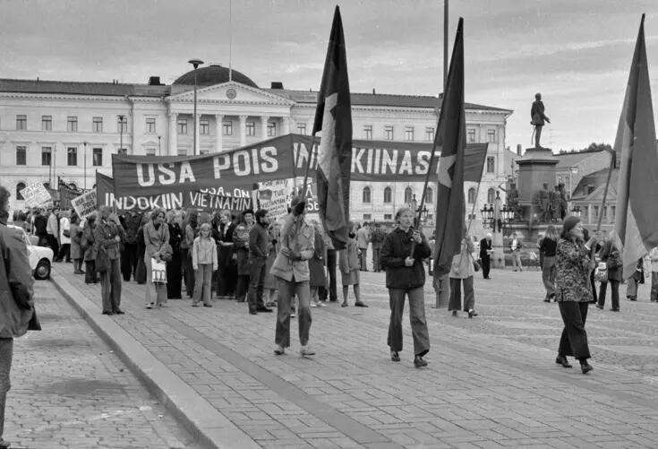 Nuorista muodostunut kulkue kulkee Senaatintorin laitaa valtioneuvoston linnalta poispäin. Kulkueen edessä kulkee kolme lipunkantajaa. Kulkueeseen kuuluu useita isoja banderolleja, joissa lukee mm. "USA pois Indokiinasta" ja "Tue Vietnamin kansan taistelua"