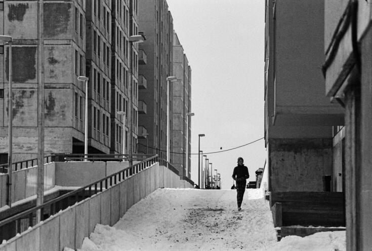En man går med händerna i fickorna i ett vintrigt Östra Böle på en gångbro mellan betonghus. 
