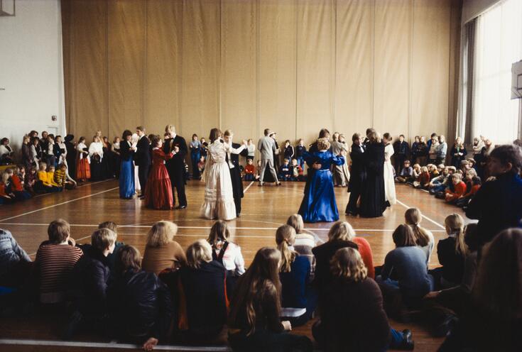 Vanhojen tanssit käynnissä koulun juhlasalissa. Tanssilattialla on vajaa kymmenkunta juhla-asuista paria. Koulun nuoremmat oppilaat istuvat ringissä tanssilattian ympärillä.