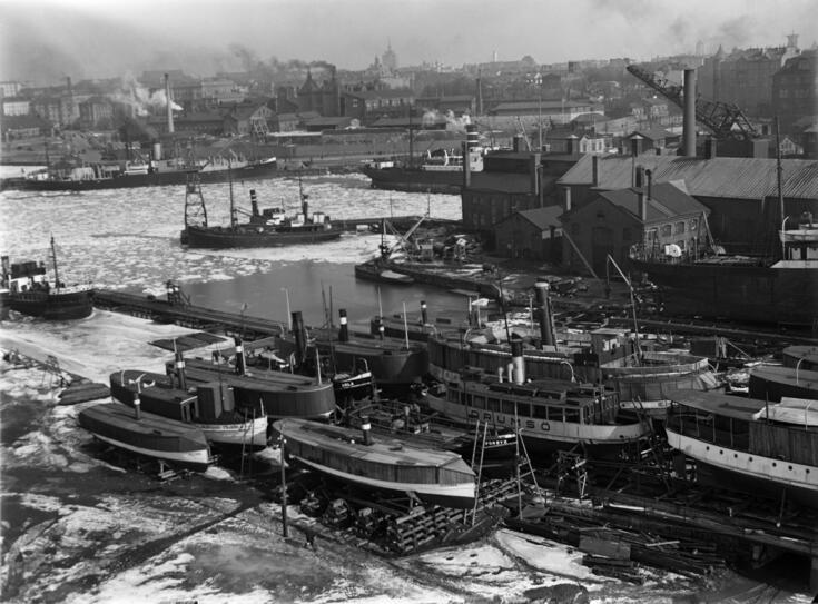 Laivoja talviteloilla Hietalahden telakalla. Taustalla savupiippujen koristamaa tiivistä kaupunkimaisemaa, Hietalahden tori ja Sinebrychoffin panimo.