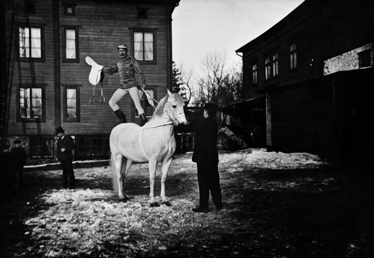 Sirkustaiteilija tasapainoilee seisaallaan valkoisen hevosen selässä Siltasaaren Sirkusmaneesin sisäpihalla.