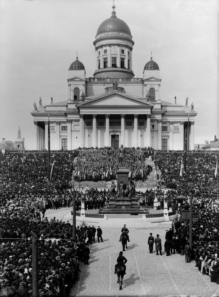 Aleksanteri II patsas sankan väkijoukon ympärillä. Patsasta lähestyy kaksi ratsastajaa edestäpäin. Taustalla näkyy Nikolainkirkko