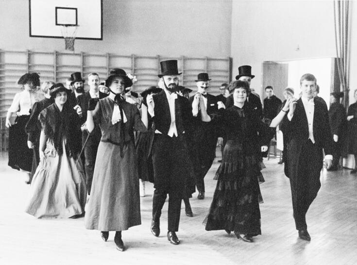 Vanhat tanssivat poloneesia. Pojat ovat pukeutuneet frakkeihin, tytöillä yksinkertaisempia mekkoja ja hatut. Osalla pojista on päässään silinteri ja tekoparrat.