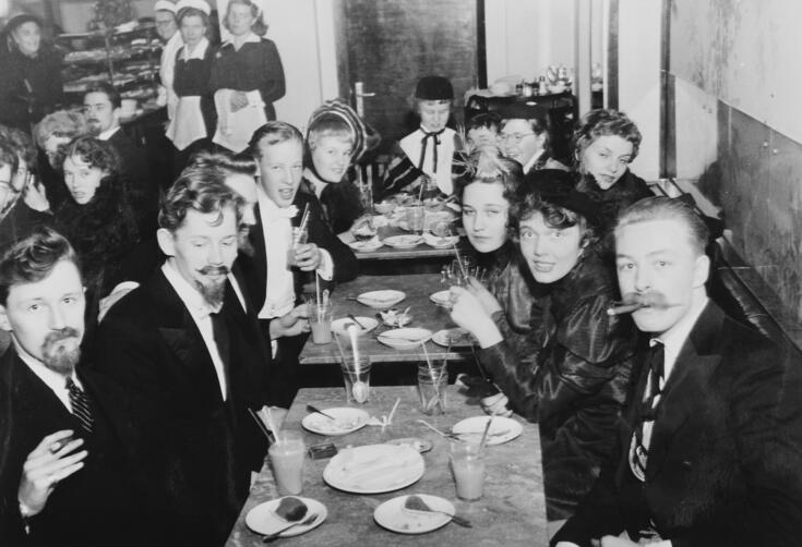 Vanhojen päivää viettäviä nuoria Fazerin kahvilassa. Etualalla istuvilla pojilla on tekoviiksiä ja -partoja, kaksi poikaa polttelee sikaria.