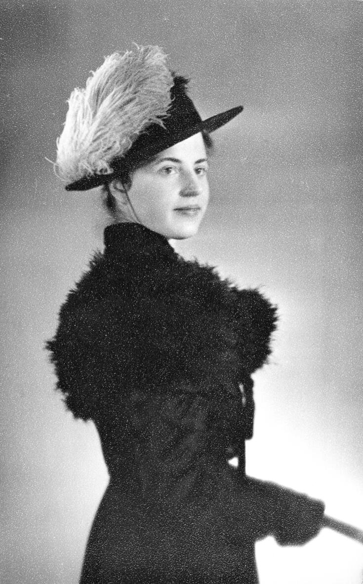 Naisopiskelija on pukeutunut vanhojen päivää varten tummaan koristeelliseen mekkoon. Puvussa on korkea kaulus ja ilmeisesti turkista olkapäillä. Tummassa lierihatussa on suuri vaalea koriste.