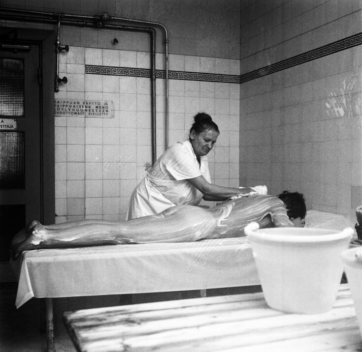 En tvätterska klädd i vit rock tvålar in ryggen på en liggande man. I bakgrunden syns vitt kakel och en skylt med text på finska: Användning av tvål och inträde i bastun som tvålig ovillkorligt förbjudet. 
