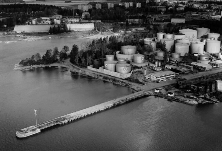 Rannalla näkyy suuri rypäs erikokoisia öljysäiliöitä, joiden kylkiin on merkitty kirjaimet TB. Ympärillä näkyy myös muita teollisuusrakennuksia. Rannasta lähtee hyvin pitkä laituri. Taustalla kauempana näkyy metsästä nousevia asuinkerrostaloja.