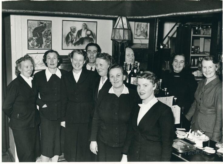 Kahdeksan naista ja kolme miestä ravintolan tarjoilutiskin edessä ja takana. Naisilla on tummat jakkupuvut ja valkoiset kauluspaidat. Seinällä on taideteoksia. 