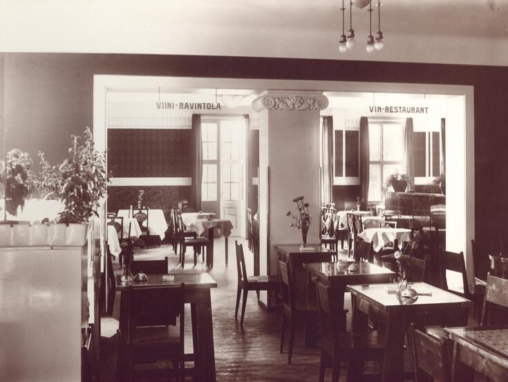 Restauranginteriör med bord och stolar. Genom balkongens glasdörrar strömmar ljus in. På borden står blommor i vaser.