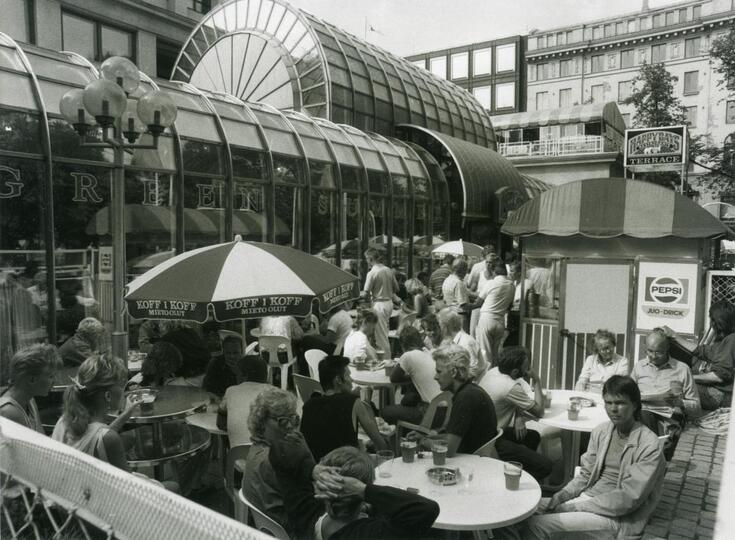 En uteservering bakom Svenska teatern. Kunder sitter vid runda bord, en del under parasoller. Restaurangbyggnaden syns i bakgrunden.