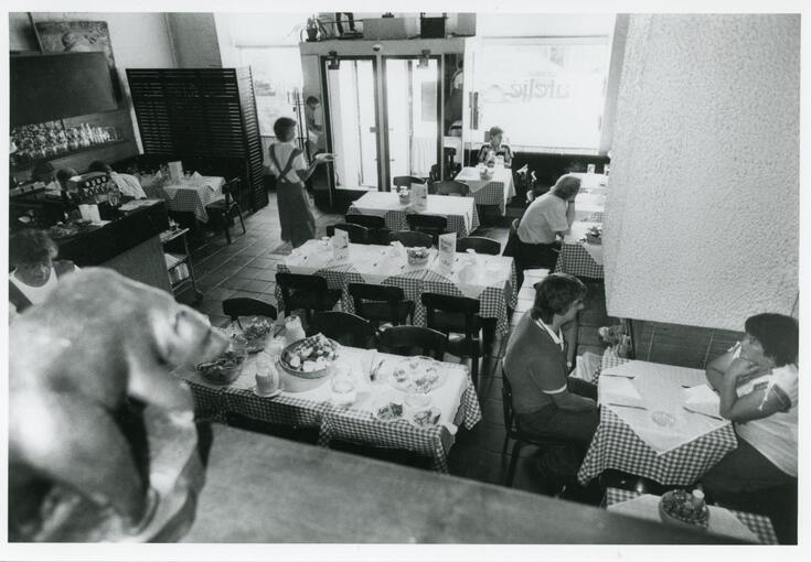 En restaurangsal under lunchtid. Kunder sitter vid bord och äter, en servitris bär på tallrikar.