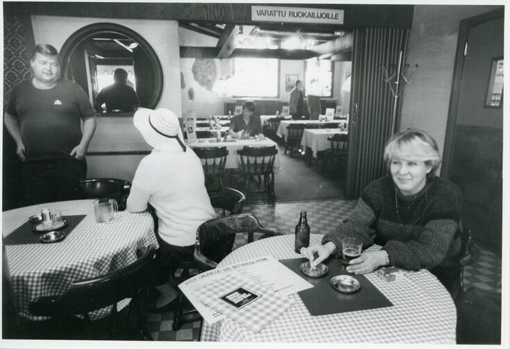 Till vänster står en man och pratar med en kvinna som sitter vid ett runt bord med ryggen mot fotografen. Hon har en vit hatt och en vit skjorta. Till höger står ett likadant bord med rutig bordduk. Vid bordet sitter en blond kvinna klädd i en mörk ylletröja. Hon håller vänstra handen runt ett ölstop och med högra handen håller hon på att fimpa en cigarrett. I bakgrunden syns en sal, ovanför dess ingång hänger en skylt med texten VAIN RUOKAILIJOILLE.