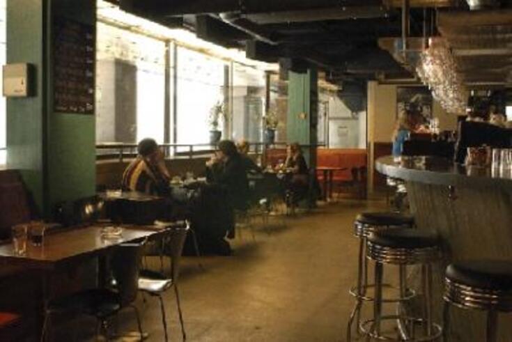 Interiör från en bar. Till vänster är fönsterväggen, till höger bardisken. Kunder sitter vid små bord intill fönsterväggen. 