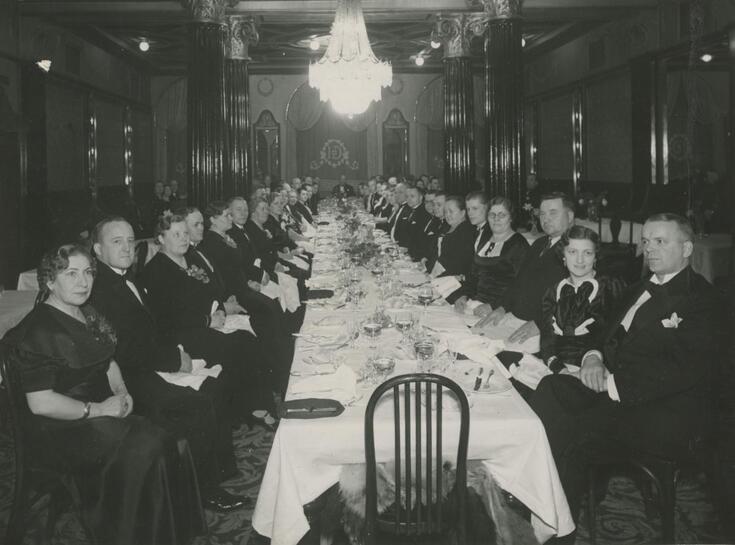 Pitkän katetun pöydän ääressä istuu juhlapukuisia miehiä ja naisia. Katossa on kristallikruunut.