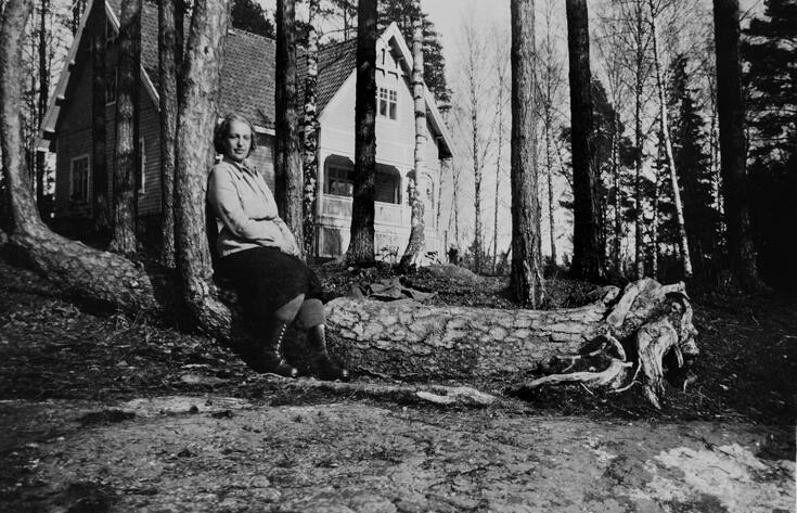 Nainen istuu puunrungon päällä, taustallaan vaalea huvila.