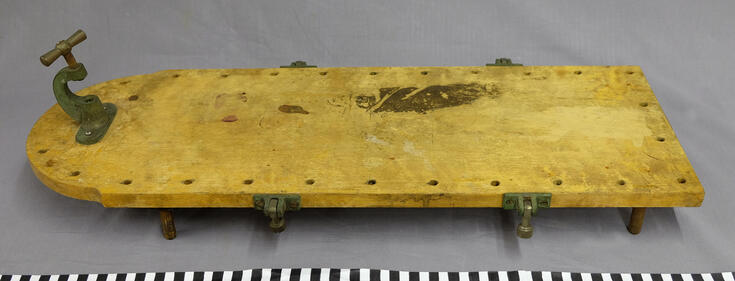 Vaaleaa puuta oleva leikkuulauta, jonka alla on matalat jalat ja metallikiinnikkeet sivuilla. Sitä on pidetty tavallisen pöydän päällä. 