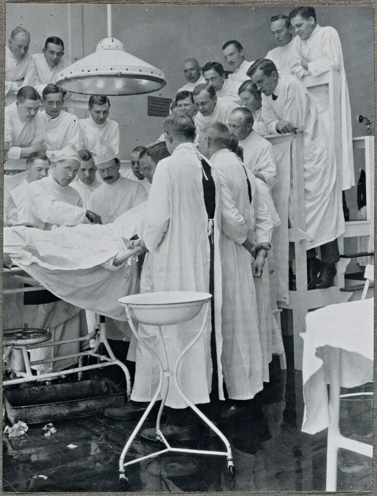 Parikymmentä henkilöä, ilmeisesti lääketieteen opiskelijaa, seuraa leikkausta leikkauspöydän ympärillä. Valtaosa heistä seisoo porrastettujen penkkirivien päällä.