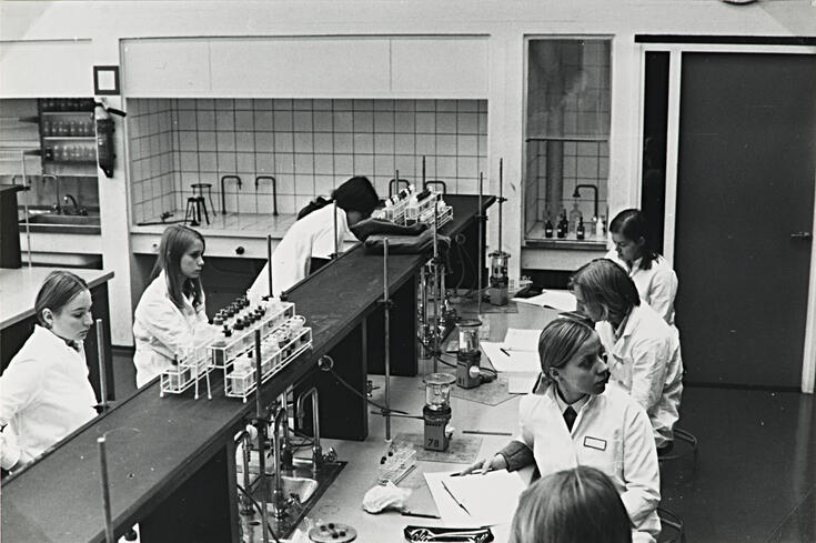 Seitsemän valkoisiin takkeihin pukeutunutta opiskelijaa istuu laboratorion työpöytien ääressä. Pöydillä näkyy laboratoriovälineitä.