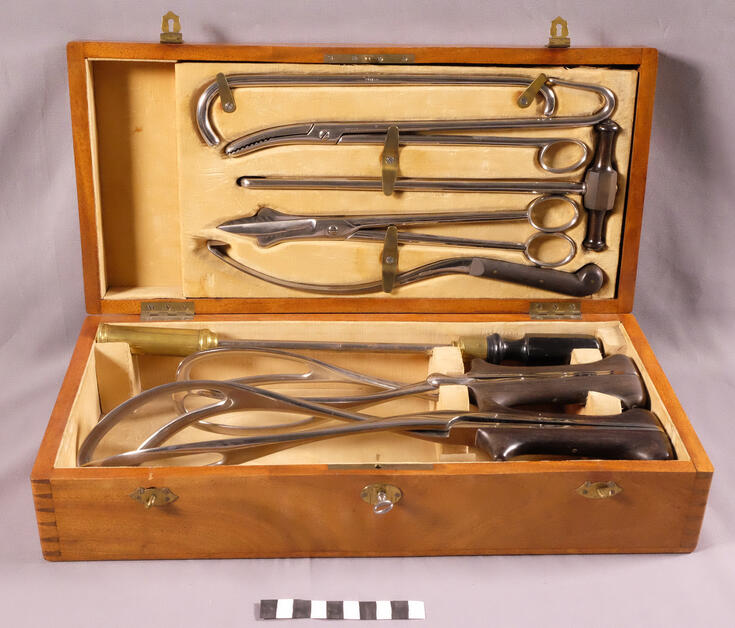 Erilaisia synnytyksessä tarvittavia metalli-instrumentteja puisessa, kellertävällä sametilla vuoratussa laatikossa. Laatikossa on lukko ja siinä pieni avain.