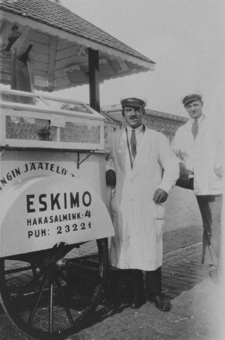 På försäljningsvagnen står det med stora bokstäver Helsingin jäätelötehdas Eskimo Hakasalmenk. 4 Puh: 23221. Intill vagnen står två män med vit rock och skärmmössa.