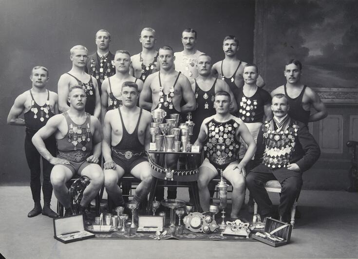 En grupp av idrottssällskapet Jyrys arbetaridrottare poserar på rad. Männen är klädda i brottartrikåer, och flera av dem har trikåerna fulla av medaljer och utmärkelser. Mitt i gruppen står ett bord som är fullt av pokaler.