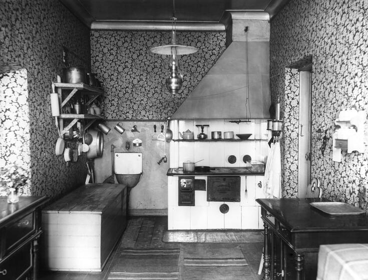 Ett kök med vedspis, liten vask och vedkista. I taket hänger en fotogenlampa.