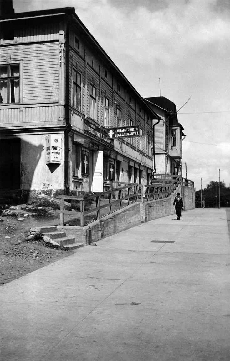 Kaksikerroksinen puutalo. Alin kerros on kivijalkaa, seinässä liikekylttejä. Jalkakäytävällä kävelee nainen.