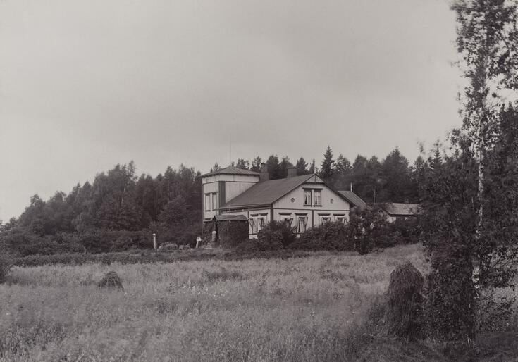 Mustavalkoisessa valokuvassa on peltomaiseman keskellä kaksikerroksinen puurakennuskokonaisuus.