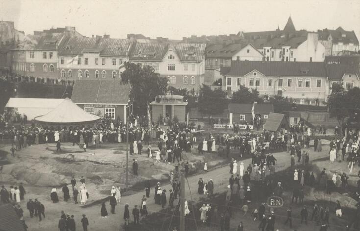 Kuvassa etualalla messualuetta ja messuvieraita, takana kaupungin puisia asuintaloja ja kivisiä kerrostaloja.