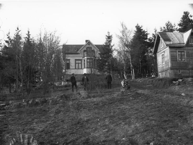 Kaksi 1,5-kerroksista puutaloa, joista toinen näkyy puoliksi, puita ja peltoa. Tilan asukkaita talon edessä. 