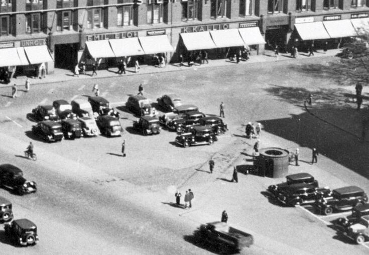 Ilmakuvan keskellä on lieriön mallinen pisoaari. Ympärillä on pysäköityjä autoja ja jalankulkijoita. Kuvan yläreunassa on Mikonkadun liiketaloja.