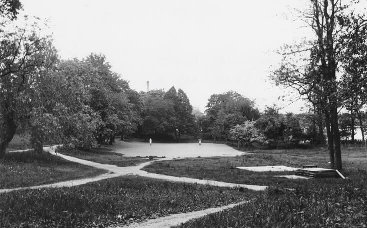 Park som korsas av grusbelagda stigar. I mitten finns ett fält med grus som kantas av träd, och där syns några människor.