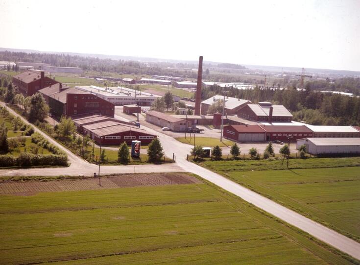 Matalalta otettu ilmakuva, jossa etualalla peltoa ja maantie, taempana tehdasrakennuksia.