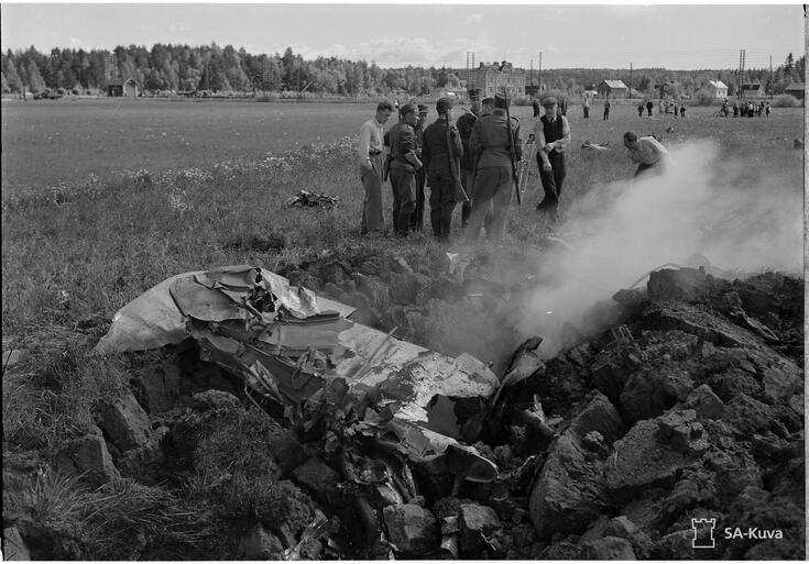 Etualalla savuavat pommikoneen jäännökset pellolla. Keskellä on ryhmä miehiä, joista osa on asepuvussa. Taustalla on Puistolan metsää ja taloja. 