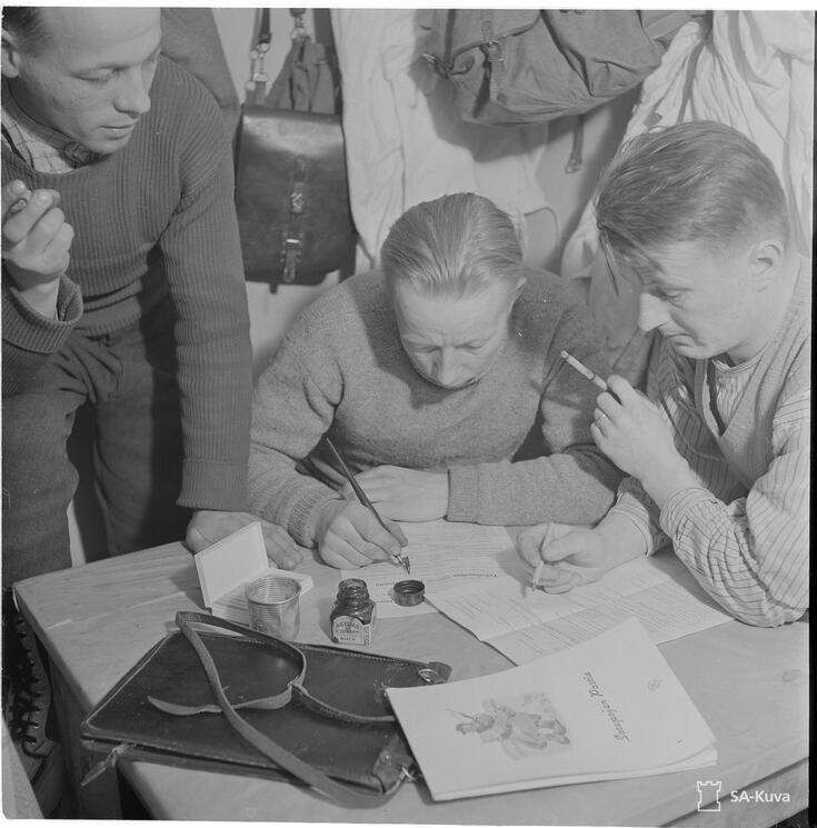 Tre rökande män fyller i skattedeklarationer vid ett litet bord. I bakgrunden hänger ryggsäckar och vita soldatdräkter.