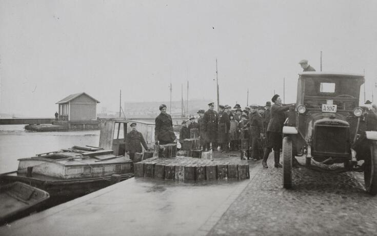 Satama-altaassa on vene, jossa seisoo mies. Altaan reunalla on kymmeniä miehiä, kuorma-auto ja kymmeniä metallikanistereita. 