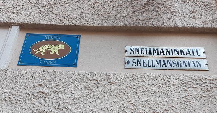 Rapattua talon seinää, johon on kiinnitetty kilpi, jossa on piirretty kuva ja teksti TIIKERI TIGERN. Sen vieressä on katukyltit SNELLMANINKATU SNELLMANSGATAN.