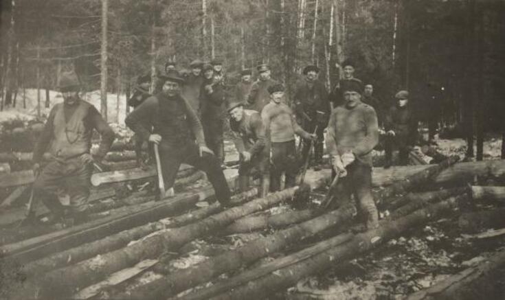 Ryhmä miehiä poseeraa kaadettujen ja oksittujen puunrunkojen ääressä metsässä.