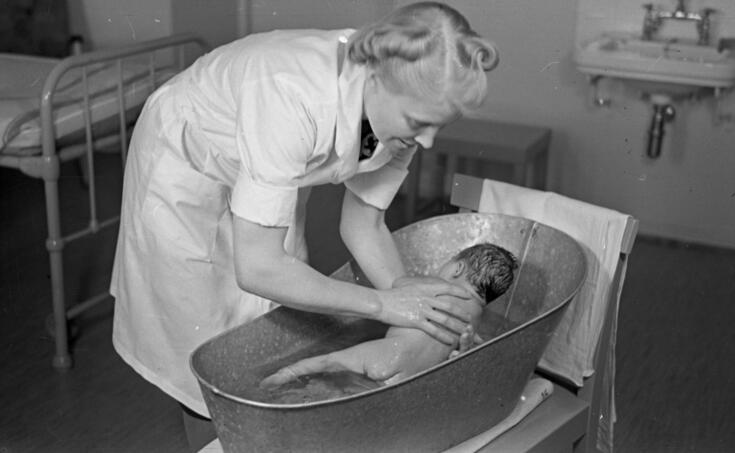 Valkoiseen mekkoon pukeutunut nainen kylvettää vauvaa peltiammeessa. Taustalla näkyy sänky ja pesuallas. 