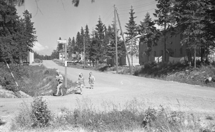 Kolme lasta kävelee maantiellä tienhaarassa kesällä. Heidän takanaan on tienviitta, jossa on kadunnimikilpi suomeksi ja ruotsiksi. Taustalla on puolitoistakerroksisia harjakattoisia taloja. 