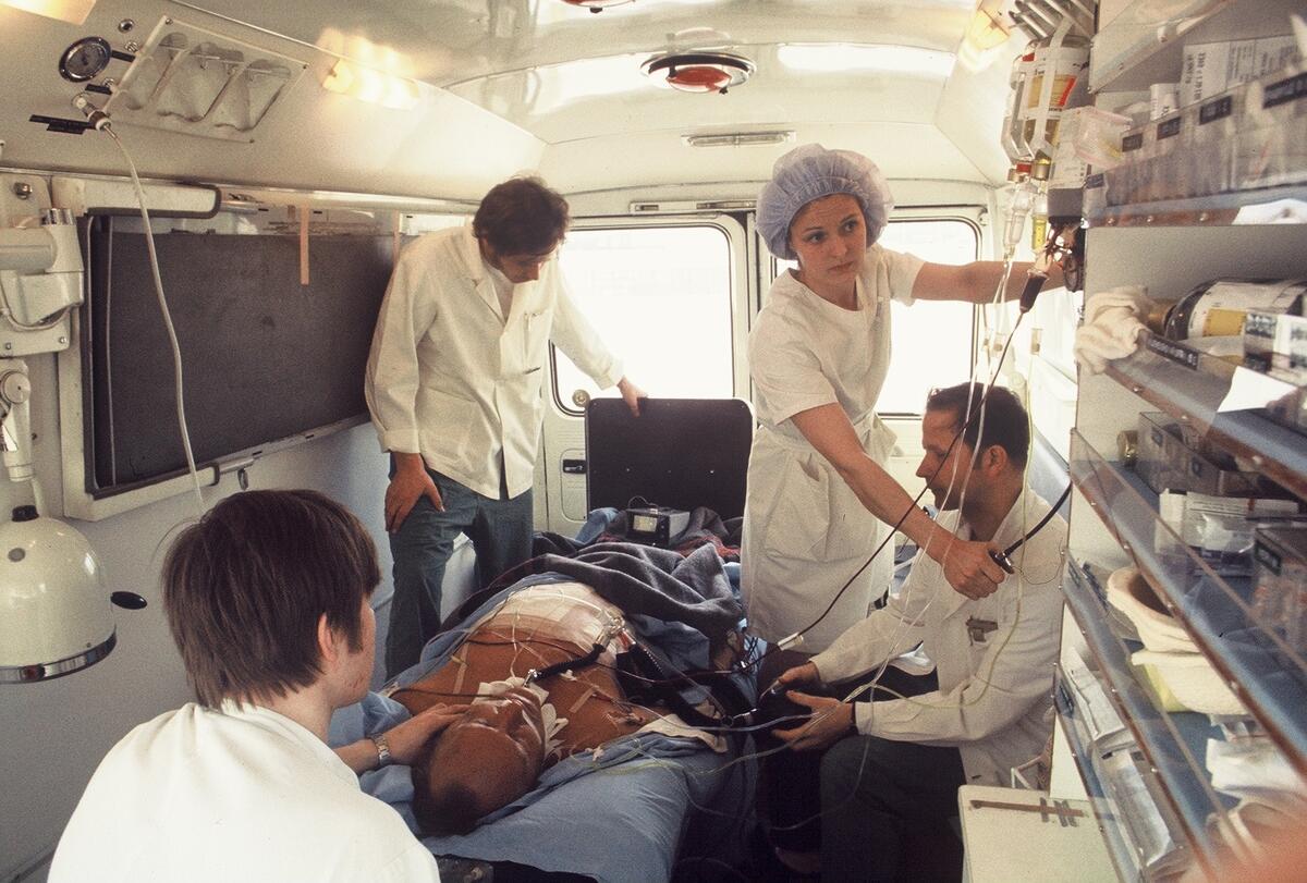 Toimenpidehuoneeksi muutettu ambulanssin sisätila. Keskellä makaa potilas, jota hoitaa neljä ihmistä.