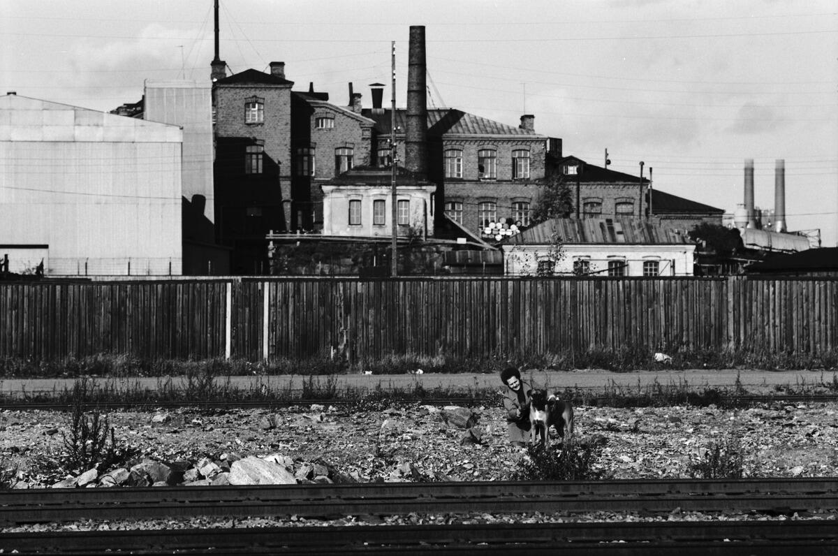 Utsikt mot norr från Skatuddskajen. I förgrunden syns järnvägsskenor, bakom den sitter en kvinna på huk bredvid en hund. Bakom dem syns Valmets varvsbyggnader som omges av ett plank. Kraftverket Hanasaari A:s skorstenar syns i bakgrunden.