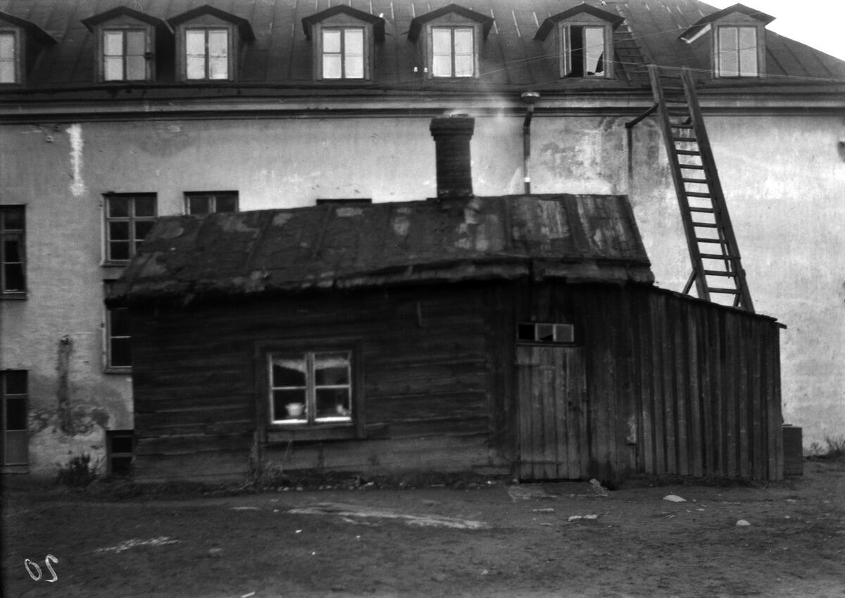 A crooked little wooden hut standing in the courtyard of a two-storey stone house.