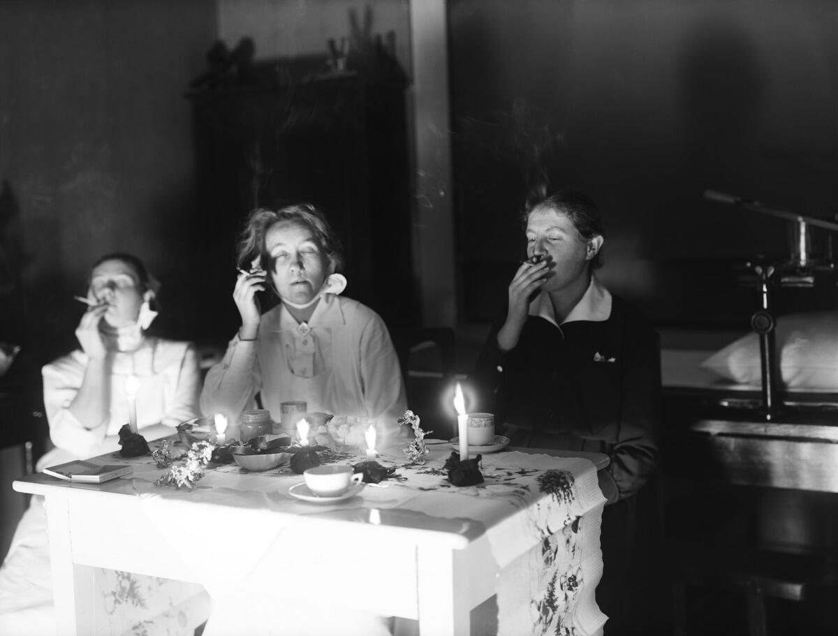 Tre sjuksköterskor sitter vid ett juldukat bord kaffebord och röker i belysning av levande ljus. På bordet syns en cigarrettask, stöpt tenn och en bordduk med julmotiv.